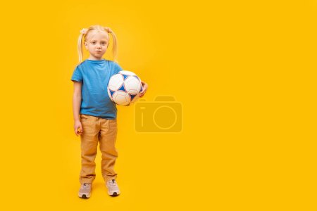 Full-length portrait of 5-6 year old child with soccer ball in isolation on yellow background. Caucasian girl with blonde hair loves football