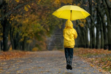 Téléchargez les photos : Enfant vole ou saute avec un grand parapluie jaune. Un enfant marche sous la pluie dans un parc d'automne. Vue arrière. - en image libre de droit