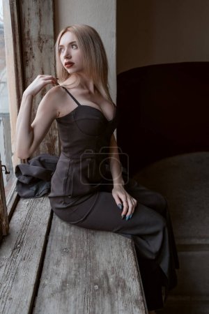 Attraktive junge Frau im schwarzen Outfit mit tiefem Dekolleté sitzt auf einem alten Fenster und blickt nach draußen. Trauriges Mädchen.