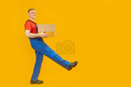 Foto de Un repartidor de paquetes sonriente que lleva la caja aislada sobre fondo amarillo. El mensajero con el paquete da un paso. Retrato de cuerpo entero. Copiar espacio, burlarse - Imagen libre de derechos