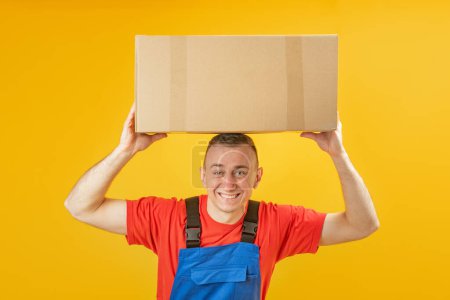 Téléchargez les photos : Jeune homme caucasien tenant une boîte sous sa tête et souriant. Courier ou chargeur tient la livraison de carton. Copier l'espace, maquiller - en image libre de droit