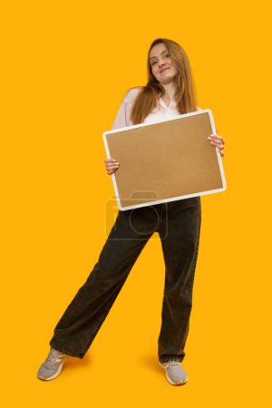 Téléchargez les photos : Jeune femme tient le carton de liège et sourire. Portrait complet en studio sur fond jaune. Espace de copie - en image libre de droit