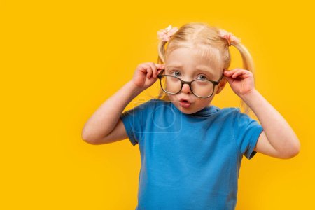 Téléchargez les photos : Surprise fille de 4 ans ajuste ses lunettes. Portrait de petite fille blonde en T-shirt bleu avec des lunettes sur fond jaune - en image libre de droit