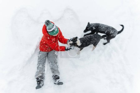 Foto de El niño se sienta en la nieve y acaricia a los perros sobre el fondo del ángel de la nieve. Concepto de perros de rescate. Vista superior - Imagen libre de derechos