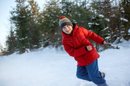 Photo for Teenage boy in red puffed jacket and knitted hat has fun in the winter in the woods. Child pulls sled - Royalty Free Image