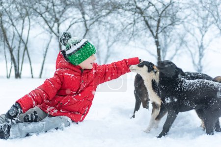 Foto de Niño en sombrero de punto se sienta en la nieve y acaricia perro mestizo. Adolescente chico juega con mascotas perros en invierno parque. - Imagen libre de derechos