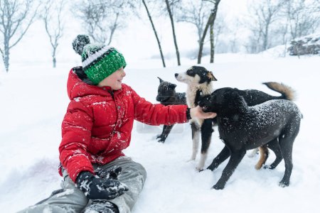 Foto de Adolescente juega con perros mascotas en el parque de invierno. Niño en sombrero de punto se sienta en la nieve y acaricia perro mestizo - Imagen libre de derechos