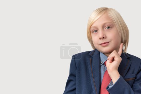 Photo for Portrait of thoughtful little boy with blond hair in blue jacket and red tie, keeps finger on chin, ponders something, isolated over grey background. People, education and school concept. - Royalty Free Image