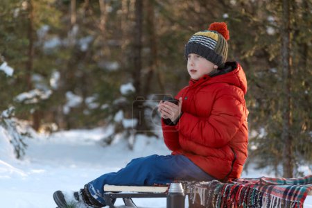 Child warms his hands on cup of hot tea in winter outside. Boy on picnic in snowy forest