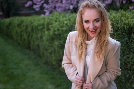 Portrait of ginger-tressed beauty in beige coat in park smiling and looking at the camera. Girl with crazy look