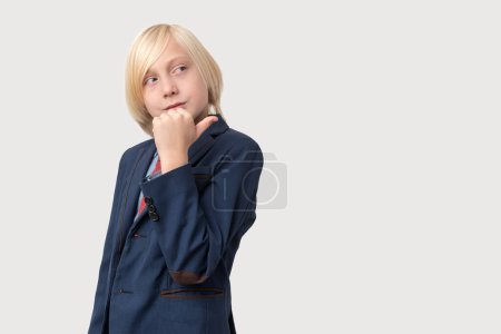 Photo for Portrait of pensive little boy with blond hair, keeps hand on chin, ponders something, dressed in dark blue suit, isolated over white background. People and business concept - Royalty Free Image