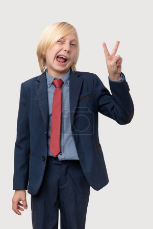 Junger schielender kaukasischer Junge mit blonden Haaren, bekleidet mit blauem Anzug und roter Krawatte, der mit beiden Händen das Siegeszeichen zeigt