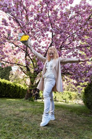 Happy middle-aged woman with bouquet of yellow tulips in blossoming tree background. Woman with flowers in spring park. Full-length portrait.