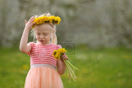 Kleines blondes Mädchen in rosa Kleid und Löwenzahnkranz mit Blumenstrauß. Sommertag. Unklarer Hintergrund
