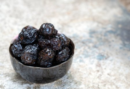 Dried Ajwa dates in a black ceramic bowl on a cement background.