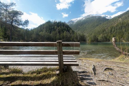 Téléchargez les photos : Banc en bois paysager au lac de montagne idyllique Fernsteinsee au Tyrol Autriche pour se détendre avec des montagnes partiellement enneigées collines et des nuages d'eau claire vert bleuâtre pause ciel et détente - en image libre de droit