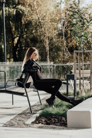 Téléchargez les photos : Jeune femme à la mode aux cheveux longs portant une combinaison monobloc à manches longues unie posant dans une veste en cuir noir et des bottes lisses sur la rue de la ville - en image libre de droit
