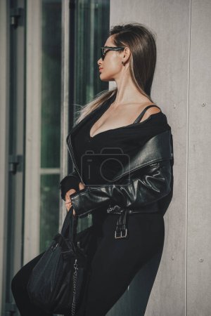 Téléchargez les photos : Jeune femme à la mode aux cheveux longs portant une combinaison monobloc à manches longues unie posant dans une veste en cuir noir et des lunettes de soleil - en image libre de droit