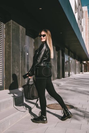 Téléchargez les photos : Jeune femme à la mode aux cheveux longs portant une combinaison monobloc à manches longues unie posant dans une veste en cuir noir et des bottes lisses sur la rue de la ville - en image libre de droit