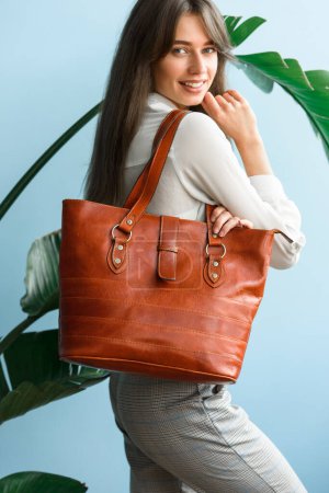 close-up photo of orange leather bag in a womans hands. indoor photo. beautiful slender girl in gray plaid pants and a white blouse posing with a handbag