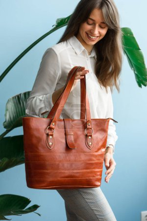 close-up photo of orange leather bag in a womans hands. indoor photo. beautiful slender girl in gray plaid pants and a white blouse posing with a handbag