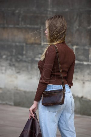 close-up photo of small brown leather bag in a woman hands. outdoors photo. Girl in a brown shirt and blue jeans