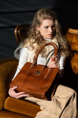 schöne Frau mit blonden Locken posiert mit einer kleinen Einkaufstasche in einem Vintage-Stuhl. Model trägt stylischen weißen Pullover und klassische Hose