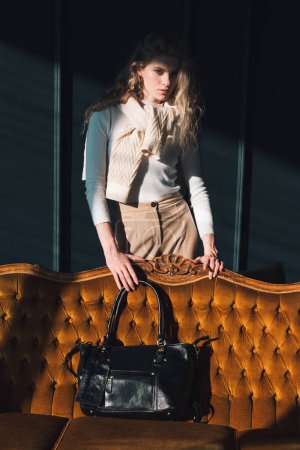 schöne Frau mit blonden Locken posiert mit einer kleinen schwarzen Einkaufstasche in der Nähe des Vintage Volor Sofas. Model trägt stylischen weißen Pullover und klassische Hose