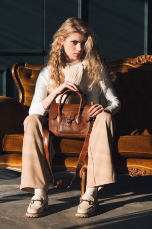 schöne Frau mit blonden Locken posiert mit einer kleinen braunen Ledertasche auf einem Vintage-Volor-Sofa. Model trägt stylischen weißen Pullover und klassische Hose