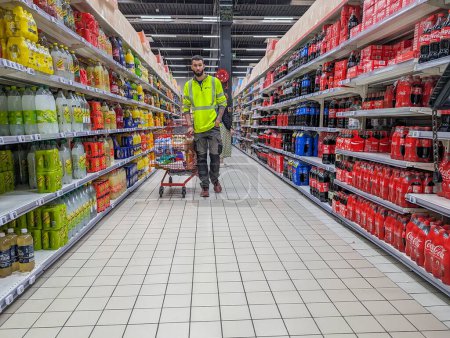 Téléchargez les photos : France, 11 avril 2024 : Homme vêtu d'une veste haute visibilité poussant un caddie dans un rayon supermarché rempli de bouteilles de boissons gazeuses. - en image libre de droit