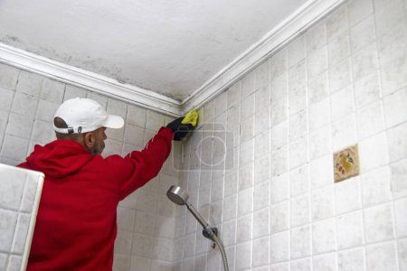 Foto de Un hombre con una sudadera roja y una gorra blanca limpiando el moho de un techo de baño. Concepto de mantenimiento higiénico. - Imagen libre de derechos