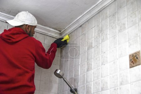 Foto de Un hombre con una sudadera roja y una gorra blanca limpiando el moho de un techo de baño. Concepto de mantenimiento higiénico. - Imagen libre de derechos