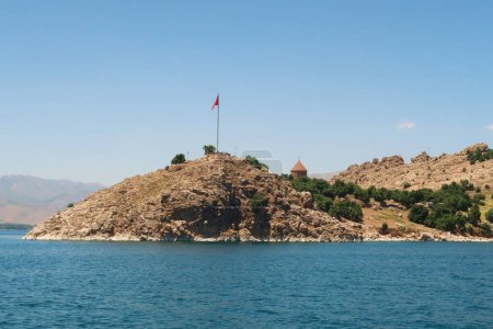 Die Insel Akdamar am Van-See, Van Golu, von einem Boot aus gesehen, die türkische Flagge weht auf einer Stange und die Spitze der armenischen Heilig-Kreuz-Kathedrale ist zu sehen, Van, Türkei 2022