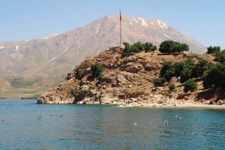 View from a boat onto Akdamar Island in the foreground with the red turkish flag on a pole and the snow covered Mount Artos in the background, Lake Van, Van Golu, Turkey 2022