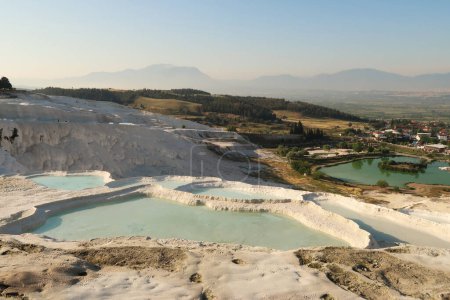 View from Pamukkale, travertine terraces filled with blue water in the front, and the entrance area at the lower gate and the town of Denizli in the back, Turkey 2022