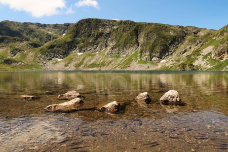 The mountains are reflecting on the smooth surface of the Kidney, Babreka Lake, one of the Seven Rila Lakes, fish are swimming in the shallow water in the foreground, Rila National Park, close to Sofia, Bulgaria 2022