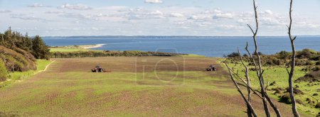 Southeastern coast of Livo island in Limfjord with tractors in agricultural field, Nordjylland, Denmark