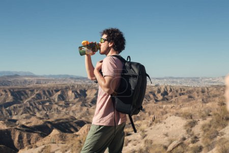 Téléchargez les photos : Prise de vue latérale du jeune homme en randonnée dans le désert et la zone chaude ou paysage. Regardez le soleil dans les lunettes de soleil de sport, boire de l'eau de voir à travers la fiole d'hydratation de l'eau, transporter un sac à dos avec matériel de randonnée - en image libre de droit