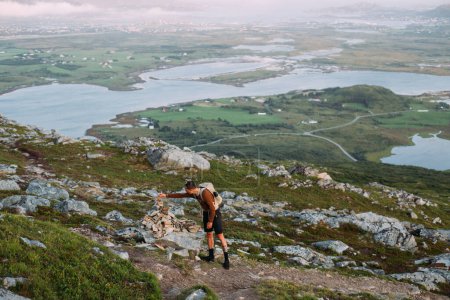 Foto de Hombre atlético en forma joven con mochila de pie en la cima de la montaña durante el atardecer o sol de medianoche en Noruega. Caminatas nocturnas de verano. Pon piedra encima de cairn. Senderismo en los países escandinavos y nórdicos - Imagen libre de derechos