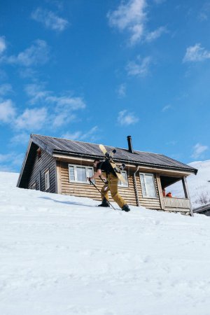 Photo for Man walk up steep ski slope to mountain cabin with help of ski poles, carry backpack with snowboard attached. Get ready for downhill on fresh powder snow. Ski or snowboard randoneering - Royalty Free Image