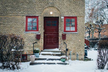 Foto de Hermoso día de invierno durante las nevadas, entrada colorida de la puerta roja brillante al edificio residencial. Estilo de vida escandinavo en la vida de la ciudad. Día de nieve en la ciudad, arquitectura escandinava. Acogedora vida - Imagen libre de derechos