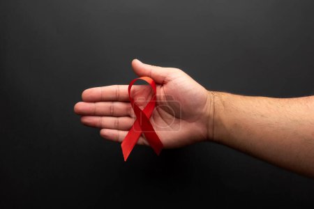 Téléchargez les photos : La paume ouverte d'un homme tenant un ruban rouge pour la Journée mondiale du sida, sur fond noir - en image libre de droit