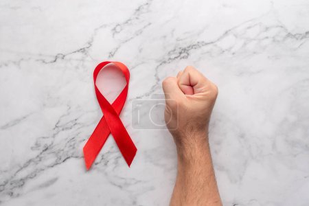 Téléchargez les photos : Un poing serré sur une table en marbre à côté d'un ruban rouge symbolisant la Journée mondiale du sida. Frappe contre les maladies du VIH. Arrêtez le sida - en image libre de droit