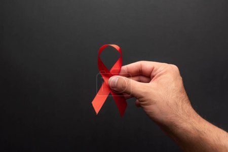 Foto per La mano di un uomo che tiene un nastro rosso a sostegno della Giornata Mondiale dell'AIDS su uno sfondo nero. dicembre 1, giorno hiv - Immagine Royalty Free
