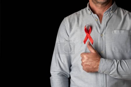 Téléchargez les photos : Un homme portant un ruban rouge de la Journée mondiale du sida sur la poitrine le pouce levé pour soutenir toutes les personnes séropositives. Lutter contre la maladie et le virus du VIH. Respect et compréhension pour les personnes vivant avec le sida - en image libre de droit