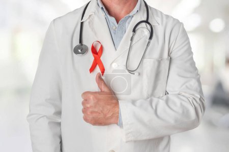 Téléchargez les photos : Médecin portant un ruban rouge sur sa poitrine, symbole de la Journée mondiale du sida, et levant le pouce pour soutenir les personnes vivant avec le VIH. Prévention et lutte contre le virus du sida - en image libre de droit