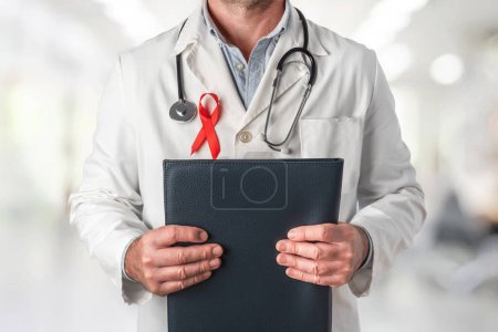 Téléchargez les photos : Docteur tenant un dossier avec des rapports médicaux et portant un ruban rouge sur sa poitrine, symbole de la Journée mondiale du sida. Soutien du personnel de santé aux personnes atteintes du VIH. Prévention et lutte contre le virus du sida - en image libre de droit