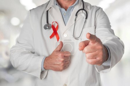 Téléchargez les photos : Docteur portant un ruban rouge sur sa poitrine, symbole de la Journée mondiale du sida. Levant le pouce d'une main et pointant vers l'avant de l'autre, indiquant un soutien et une compréhension pour toutes les personnes infectées par le virus du VIH - en image libre de droit