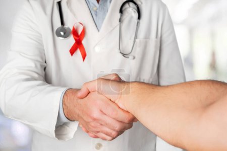 Téléchargez les photos : Un médecin portant un ruban rouge sur la poitrine, symbole de la Journée mondiale du sida, serre la main d'un patient séropositif. Soutien du personnel de santé pour toutes les personnes infectées par le VIH - en image libre de droit