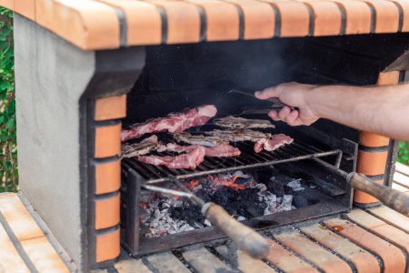 Foto de Asar carne en una barbacoa de ladrillo con brasas brillantes. Lomos de cerdo, chuletas y costillas cocinando en la parrilla - Imagen libre de derechos
